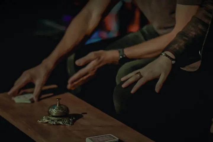Hands hovering over a table with a brass bell and a deck of cards